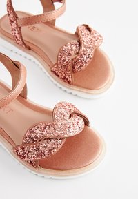Sandal z rožnatim zlatom z bleščečo prepleteno trakasto zasnovo, gladkim satenskim zaključkom in belo gumijasto podplato za oprijem.
