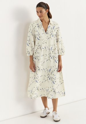 Kvinde med mørkt hår i en hestehale iført en cremefarvet blomsterprintet midi kjole med 3/4 ærmer og hvide sneakers, stående op ad en hvid væg.