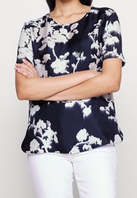 Blůza s květinovým vzorem v námořnické a krémové barvě, vyrobená z hladkého materiálu. Má kulatý výstřih a krátké rukávy, s volným střihem.