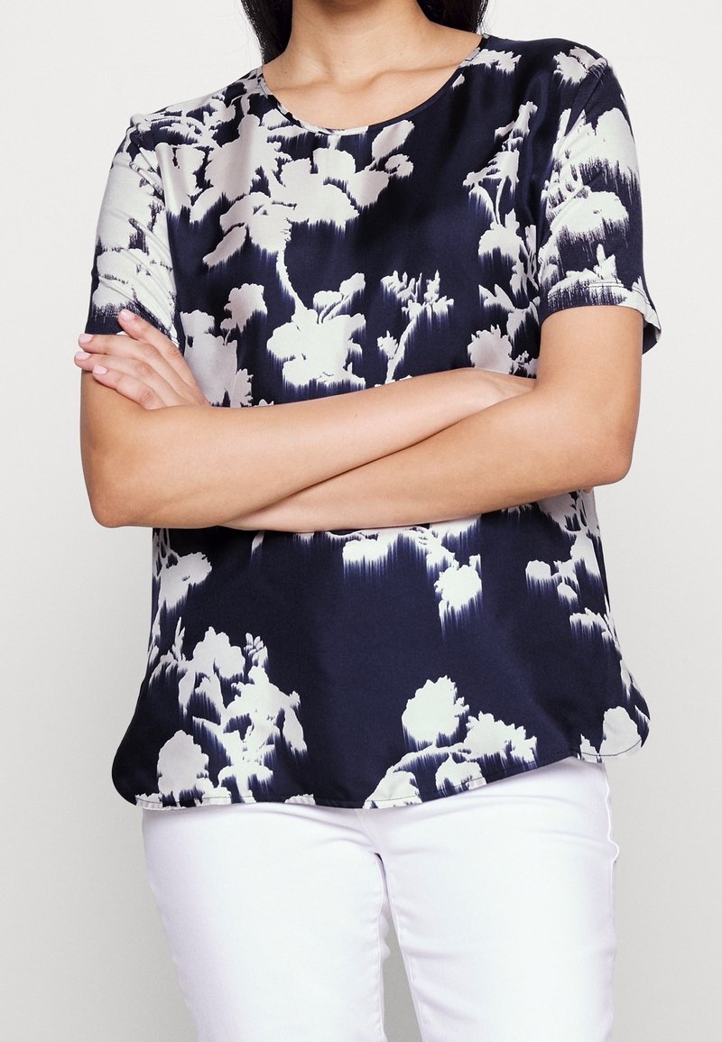 Blůza s květinovým vzorem v námořnické a krémové barvě, vyrobená z hladkého materiálu. Má kulatý výstřih a krátké rukávy, s volným střihem.