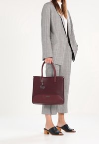 Ciemnobordowa skórzana torba typu tote o teksturowanej powierzchni, gładkim spodzie, podwójnych uchwytach i metalowej zawieszce. Noszona z szarym, kraciastym garniturem i czarnymi klapkami.