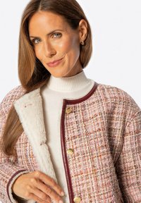 Kobieta z długimi brązowymi włosami, ubrana w różową i białą kurtkę z tweedu z złotymi guzikami oraz kremowym futerkiem na białej golfie.