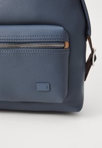 Niebieska torba z teksturowanej skóry wyposażona w przednią kieszeń na suwak oraz małą naszywkę z logo, z brązowymi akcentami i usztywnionym kształtem.