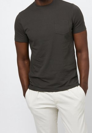 Homme portant un t-shirt gris foncé à manches courtes avec une poche poitrine et un pantalon blanc, les mains dans les poches, sur un fond uni.