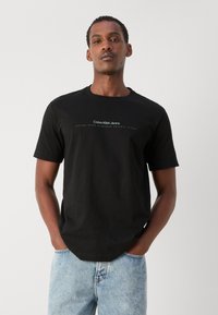 Juodas marškinėlis su apvaliu kaklu, ant kurio yra balti užrašai "Calvin Klein Jeans" ir "Įkurtas devyniasdešimt aštuoniasdešimt." Kasdienis siluetas, trumpomis rankovėmis.