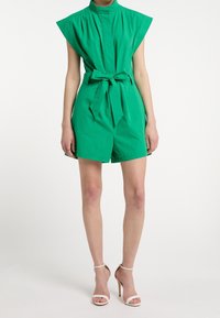 Combinaison verte en mélange de coton avec un col haut, des manches courtes et une ceinture à la taille. Style short avec une texture lisse et sans motifs visibles.