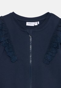 Giacca a zip blu navy con dettagli in pizzo testurizzato a volant sulla spalla sinistra e zip argentata. Materiale in cotone morbido.