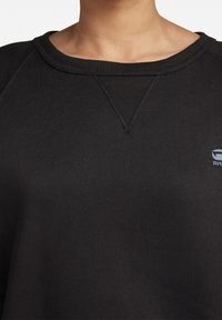Felpa nera con scollatura ampia a coste, cuciture decorative e una piccola applicazione del logo sul petto. Tessuto morbido con una vestibilità rilassata.