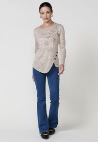 Beige Langarmtop aus strukturiertem Stoff, mit floraler Bestickung auf der linken Seite und schwarzen Knöpfen am rechten Saum. Kombiniert mit blauen Schlagjeans.