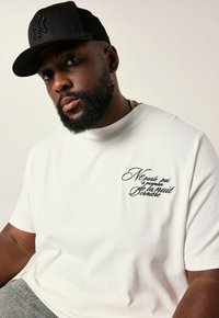 Behaarde man in een zwarte pet en een wit T-shirt met Franse tekst geborduurd op de borst, zittend tegen een effen achtergrond.