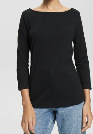 Frau trägt ein schwarzes langärmeliges Shirt mit Bootsausschnitt und umgeschlagenen Ärmeln, kombiniert mit hellblauen Jeans, steht vor einem neutralen Hintergrund.