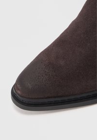 Stivale alla caviglia in suede marrone scuro con punta affilata, texture liscia e suola in gomma nera. Presenta una leggera lucentezza e cuciture ordinate lungo il bordo.
