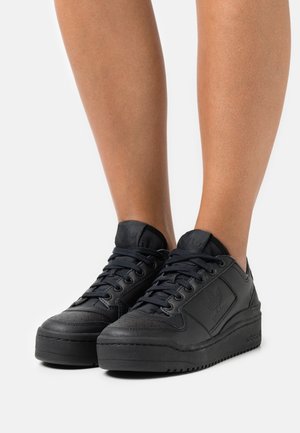 Baskets plateformes basses noires Adidas avec lacets, portées sur des jambes nues, montrant une semelle texturée et un logo embossé sur le côté et la languette.