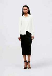 Vit blus med långa ärmar och spetsig krage, kombinerad med en svart knälång kjol. Svarta sandaletter med remmar fullbordar stilen.