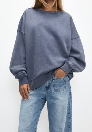 Kobieta w oversize wyblakłej niebieskiej bluzie z długimi rękawami i jasnoniebieskich spodniach jeans z wysokim stanem, z jedną ręką w kieszeni, stoi na białym tle.