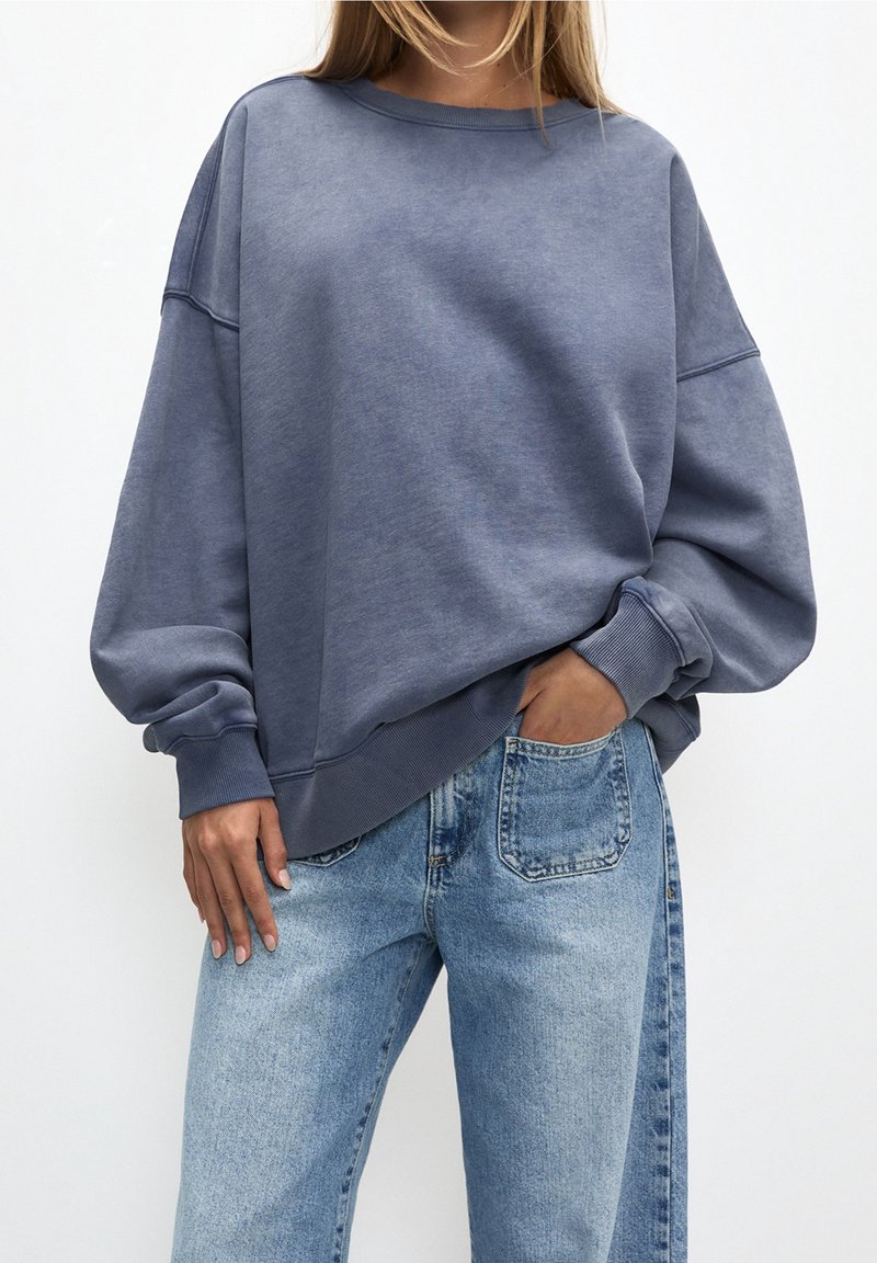 Femme portant un sweat-shirt bleu délavé oversize à manches longues et un jean taille haute bleu clair, une main dans la poche, debout sur un fond blanc.