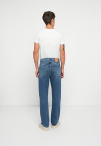 Jeans de mezclilla azul con diseño de pierna recta, cinco bolsillos y un parche de cuero marrón claro en la parte trasera de la cinturilla. Combinados con una camiseta blanca.