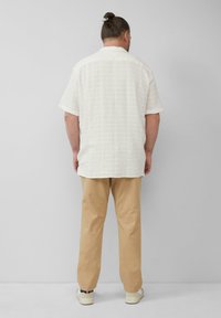 Ein Mann mit einem Dutt trägt ein weißes kurzärmliges Hemd, beige Hosen und weiße Turnschuhe und steht vor einer schlichten hellgrauen Wand.