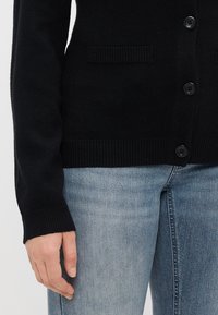 Μαύρο μάλλινο κασκόλ με ραβδωτές μύτες και μανσέτες, έξι κουμπιά και δύο μπροστινές τσέπες. Συνδυασμένο με ανοιχτό μπλε τζιν.