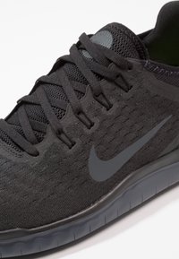 Chaussure de sport noire avec tige en mesh respirant, lacets plats et un swoosh Nike proéminent. Présente une surface texturée et une semelle en caoutchouc flexible.