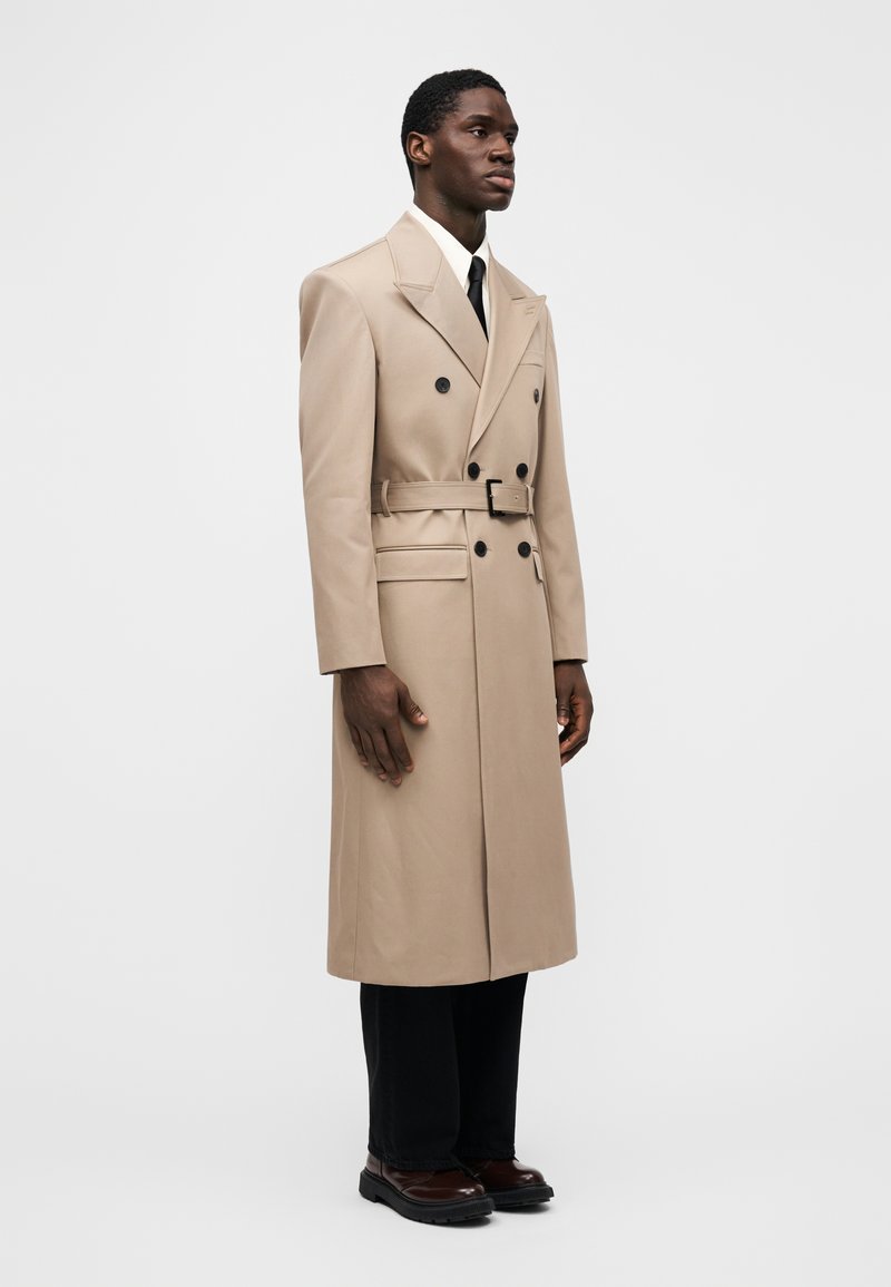Trench-coat beige à double boutonnage avec une ceinture à la taille, de larges revers et des boutons noirs. Stylé avec un pantalon noir et des chaussures marron.