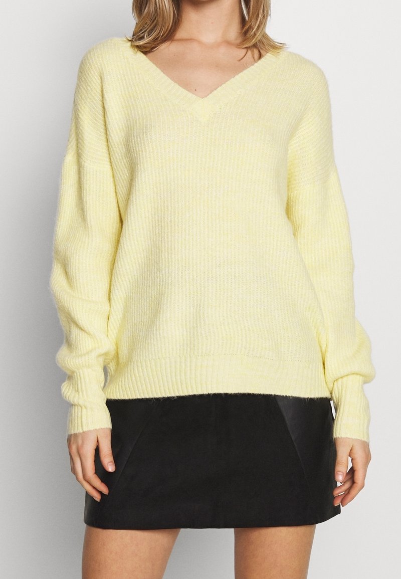 Hellgelbes, übergroßes V-Ausschnitt-Pullover mit geripptem Design und langen Ärmeln, kombiniert mit einem schwarzen, knielangen Rock aus Kunstleder.