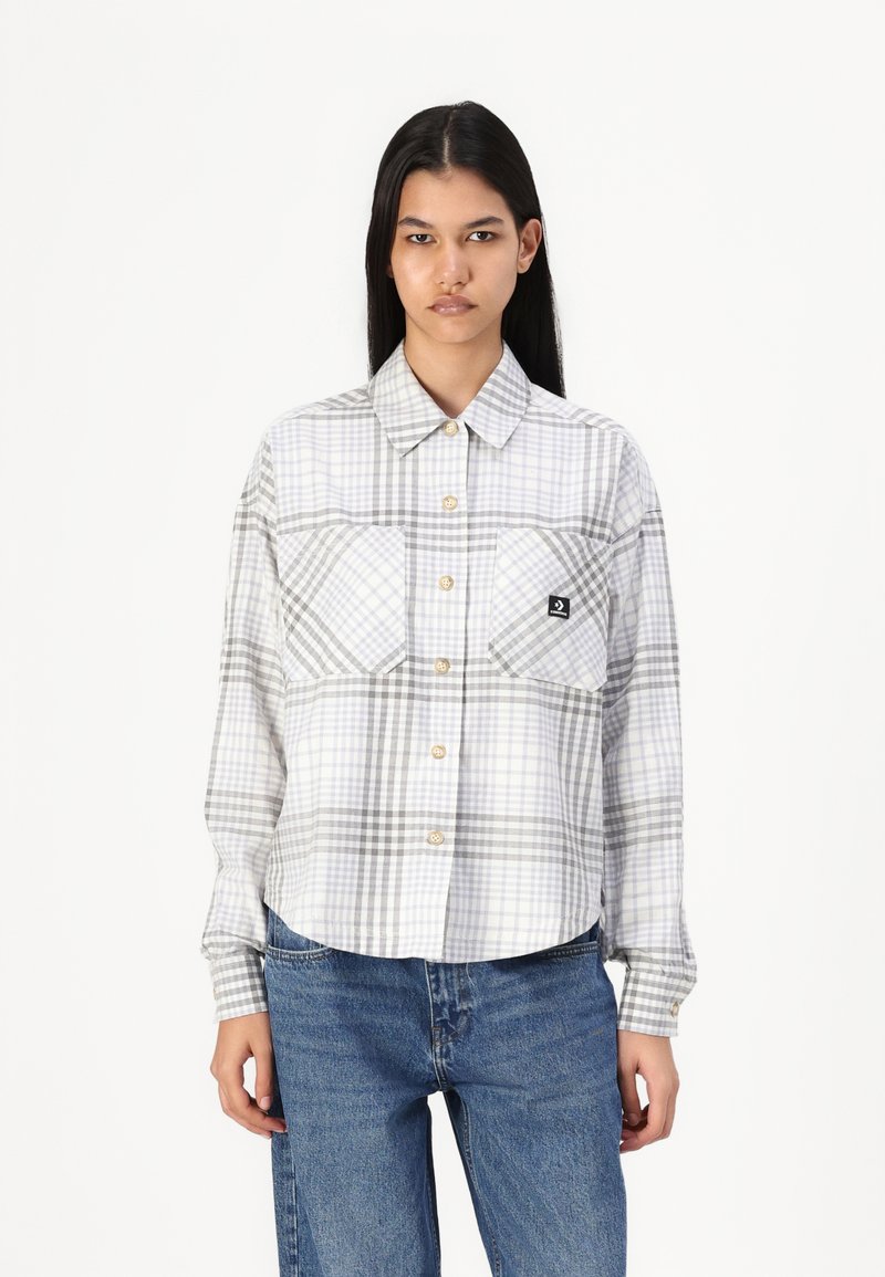 Camisa de botão em padrão xadrez branco e cinza, com dois bolsos frontais e mangas longas, feita de um tecido leve. Possui botões castanhos na pala.