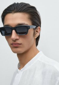 Black rectangular sunglasses with dark lenses; plastic frame; worn with a white linen garment. Lightly tousled dark hair.