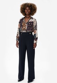 Het overhemd met lange mouwen heeft een veelkleurig bloemen- en geometrisch patroon; gecombineerd met hoge, wijde zwarte broeken en strakke zwarte schoenen.