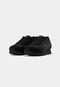 Adidași negri cu sistem de șireturi și talpă joasă, având logo-ul "cK" pe lateral, prezentati dintr-o perspectiva unghiulară frontală.