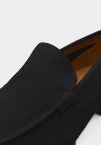 Μαύρο σουέντ ανδρικό μοκασίνι χωρίς κορδόνια με ραμμένες λεπτομέρειες και επένδυση εσωτερικά σε ταμπά χρώμα, με γωνιακή προβολή που δείχνει το πάνω μέρος και το πλάι.