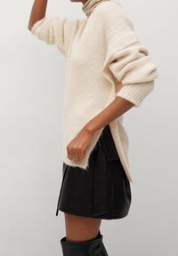 Kremowy, oversize'owy sweter z prążkowanymi rękawami, bocznymi rozcięciami i miękką fakturą. Połączony z czarną skórzaną spódnicą i wysokimi kozakiem.