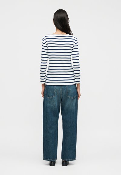 Μπλούζα μακριά μανίκια σε ναυτικό μπλε και λευκές ρίγες με λαιμόκοψη τύπου βάρκα, σε συνδυασμό με φαρδιά μπλε τζιν παντελόνια. Απλός σχεδιασμός, βαμβακερό υλικό. Πλάτη.