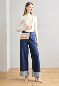 Haut long à manches crème, jeans en denim foncé à jambes larges avec des poignets gris contrastants, sac bandoulière rose avec des accessoires dorés, sandales à talons transparents.