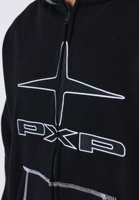 Sudadera con cremallera negra hecha de tejido suave, que presenta un logotipo bordado en blanco y un diseño geométrico en el pecho. Acentuada con costuras en contraste.