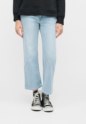 Persona che indossa jeans larghi azzurri e sneakers nere alte con lacci bianchi, in piedi davanti a uno sfondo semplice.