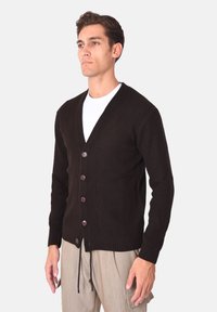 Cardigan marrone a coste con scollo a V e cinque bottoni frontali. Presenta un design aderente con un dettaglio a fiocco sull'orlo. Indossato sopra una t-shirt bianca.