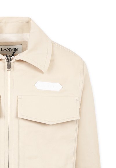 Beige utilityjack met een ritssluiting, puntige kraag, borstzakken en een getextureerde witte logo patch aan de linkerkant.