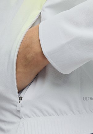 Käsi työnnetty valkoisen, teksturoidun takin sivutaskuun, jossa on osittain auki oleva vetoketju ja pitkä hiha.