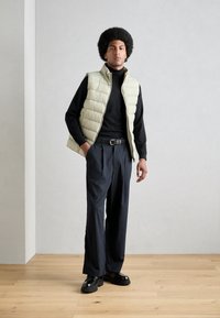 Light green puffy vest over a black turtleneck, paired with navy pinstriped wide-leg trousers and black chunky shoes on a wooden floor.