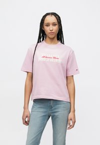 Hellrosa Baumwoll-T-Shirt mit kurzen Ärmeln, mit weißem und rotem "TOMMY HILFIGER" Schriftzug und Logo. Kombiniert mit hellblauen Jeans.
