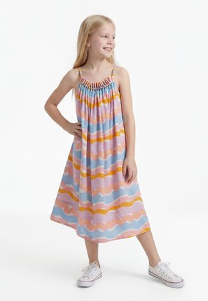 Nuori tyttö hymyilee, pukeutuneena hihattomaan värikkääseen raidalliseen mekkoon, jossa helmikoristellut olkaimet, ja valkoiset lenkkarit, seisoo käsi lanteella.