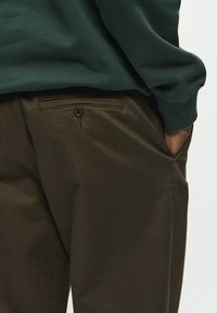 Brown cotton pants with a smooth texture, featuring a side pocket and a buttoned back pocket. Worn with a green long-sleeve top.