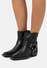 Botines de cuero negro con punta afilada, tacón de altura media y una correa decorativa con acentos metálicos. Textura suave y puño doblado.