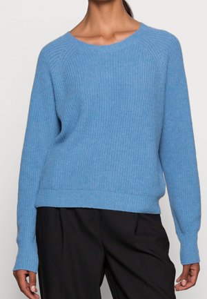 Personne portant un pull en maille bleu clair à manches longues et un pantalon noir, debout devant un fond uni.