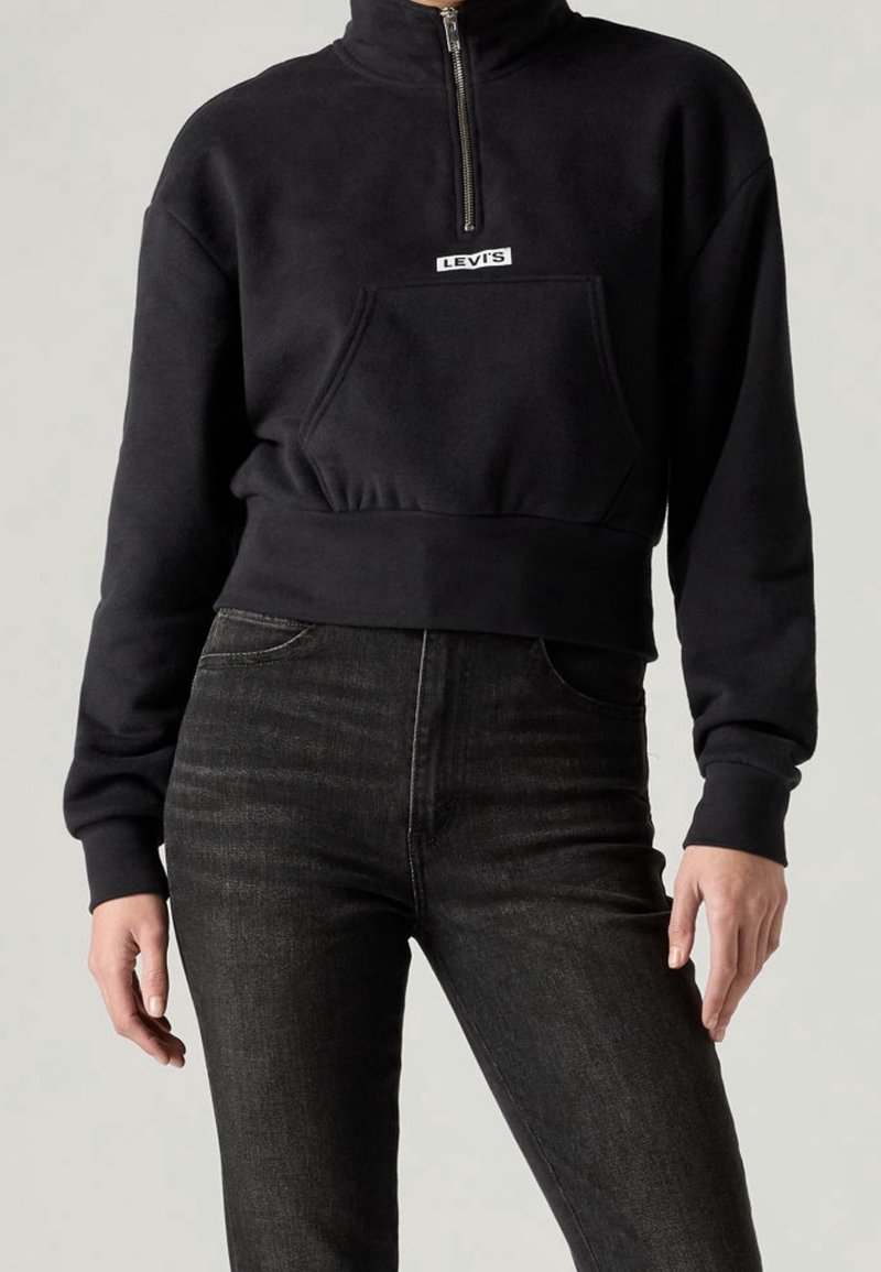 Sweat-shirt court noir avec fermeture éclair à l'avant et poche kangourou, associé à un jean taille haute gris foncé sur une personne debout.