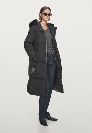Schwarze Puffjacke mit einem mit Fell gefütterten Kapuze, silbernem Reißverschluss und seitlichen Taschen. Kombiniert mit einem grauen Oberteil und dunklen Jeans sowie schwarzen Schuhen.