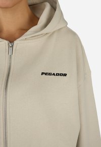 Μπεζ φούτερ με φερμουάρ και επένδυση στην κουκούλα, διαθέτει μαύρο κεντητό λογότυπο "PEGADOR" στην αριστερή πλευρά του στήθους, κατασκευασμένο από μαλακό ύφασμα.