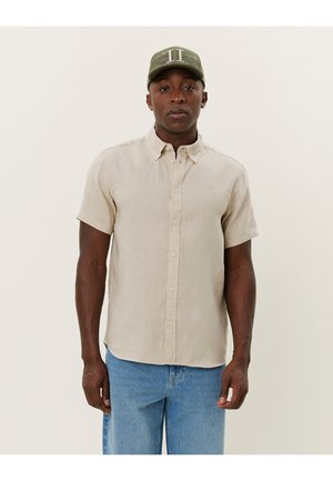 Beige Leinenhemd mit kurzen Ärmeln, Button-Down-Kragen, vorderem Knopfverschluss und leichter Textur. Kombiniert mit hellblauen Jeans und einer grünen Kappe.