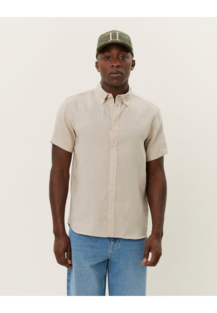 Cămașă din in bej cu mâneci scurte, cu guler tip button-down, nasturi în față și o ușoară textură. Asortată cu blugi din denim bleu deschis și o șapcă verde.
